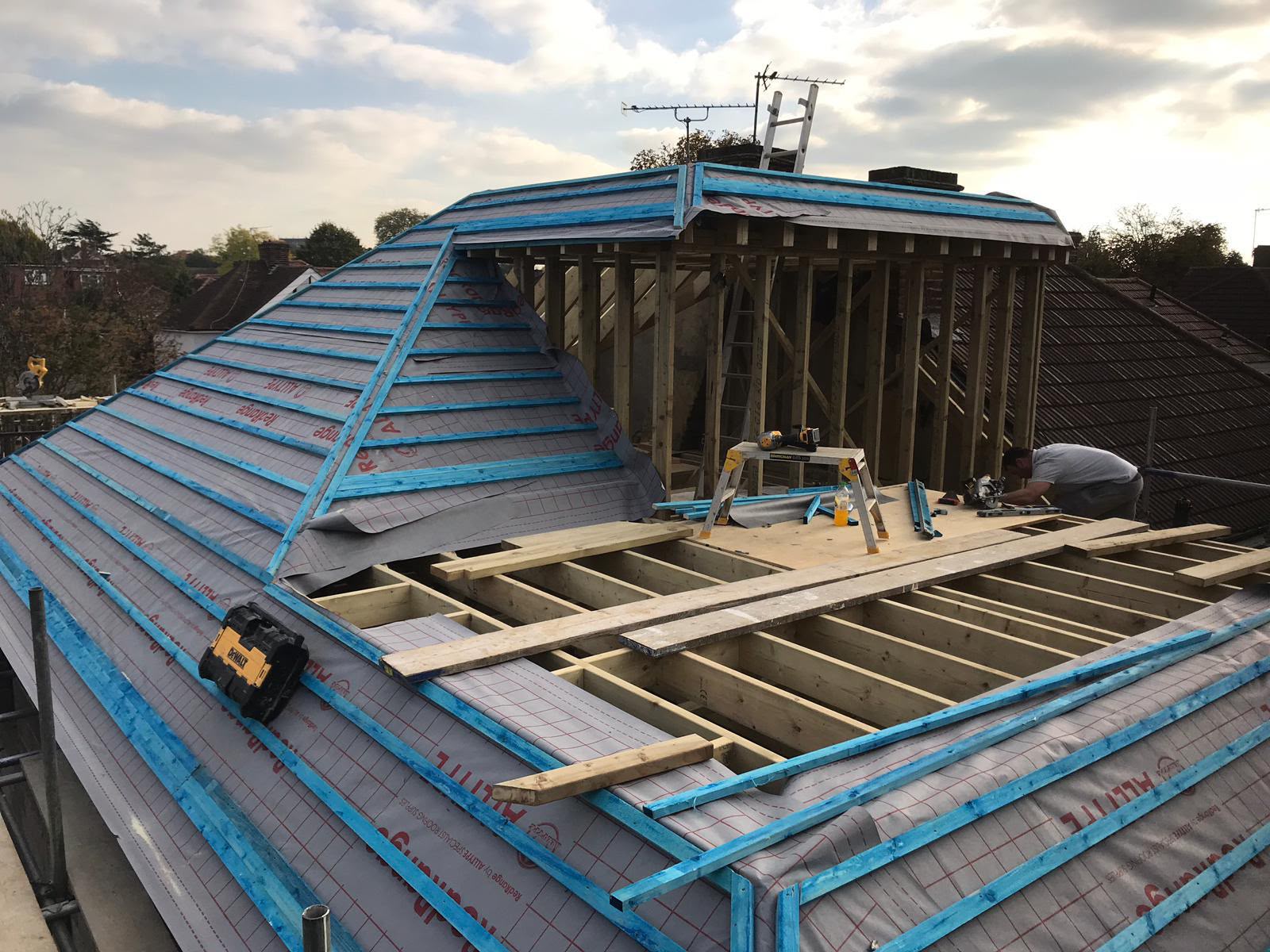 A carpenter working on a new loft floor beside a timber-framed dormer, with the hipped roof covered in membrane and battens