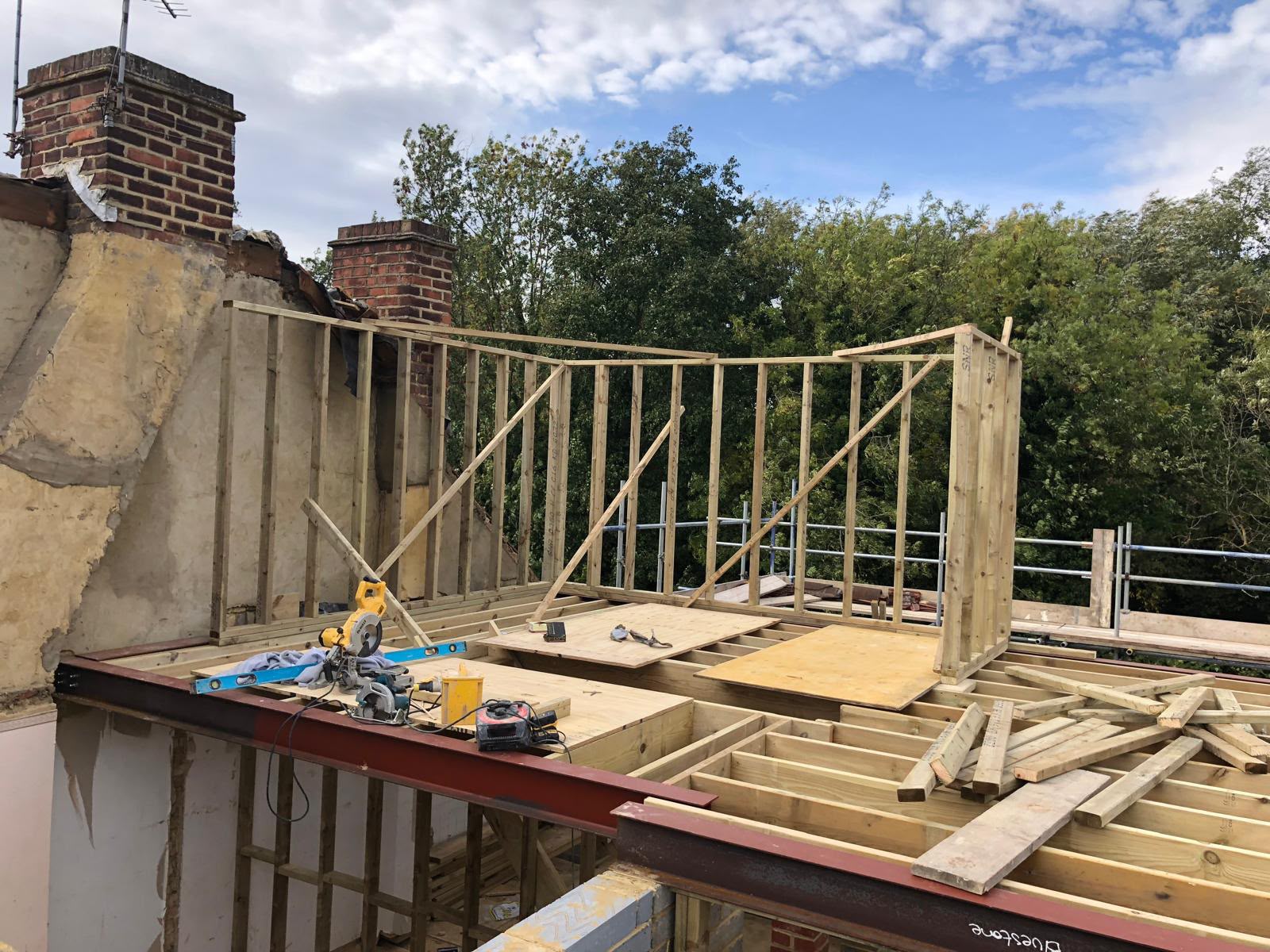Timber dormer walls framed on a new loft floor above red steel beams, beside two brick chimneys and a stripped-back roof