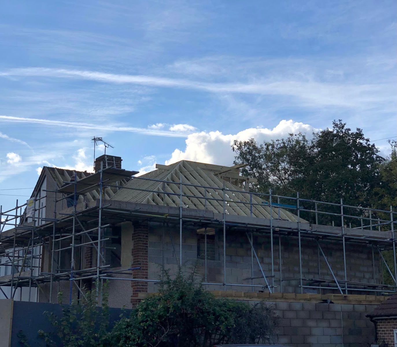 New timber roof rafters and hips standing on blockwork walls behind scaffolding, seen from the street against a blue sky
