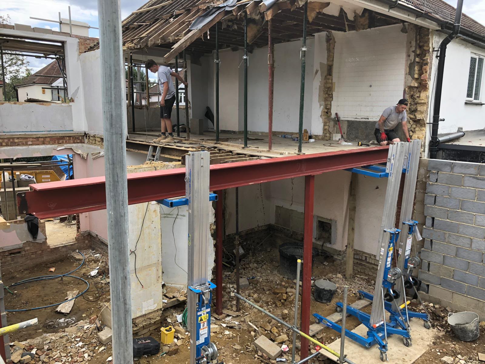 Builders lifting a red steel beam into place on material hoists at the opened-up rear of a house, with the first floor propped above
