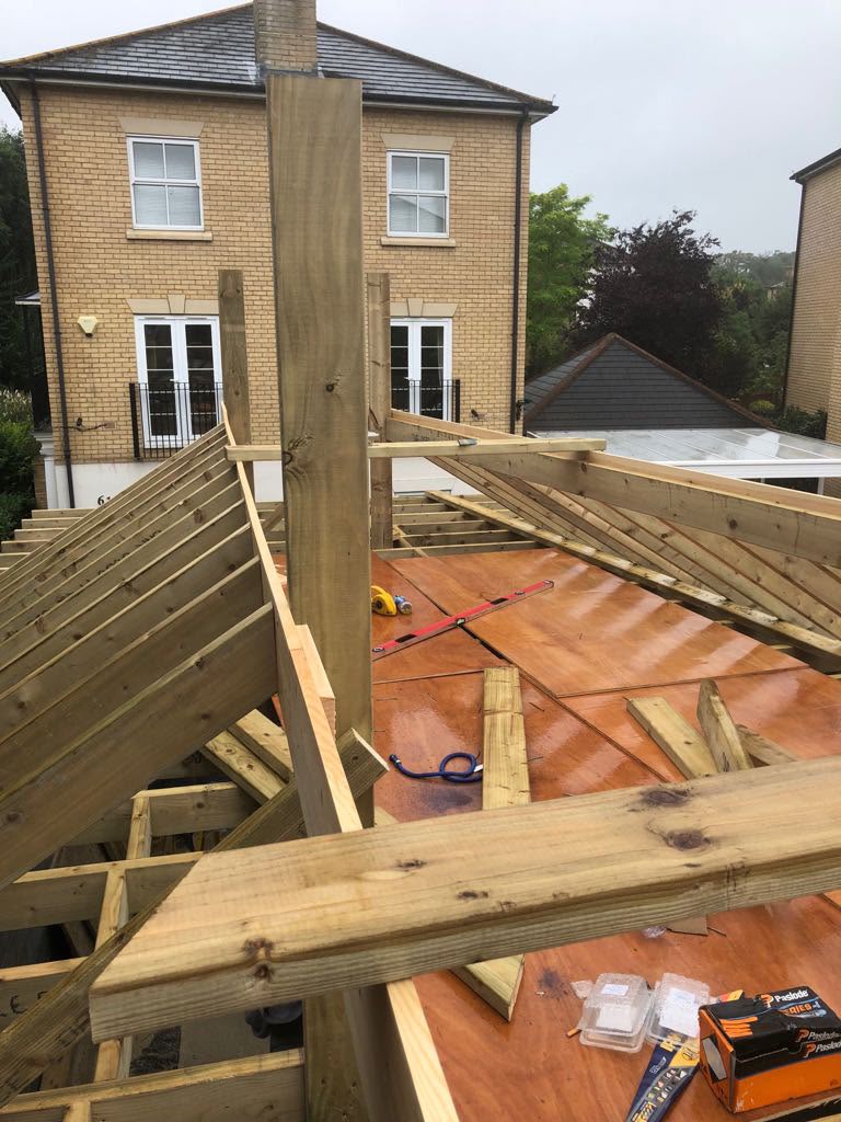 Timber hip rafters being framed over a plywood deck on a new garage, with a brick house in the background