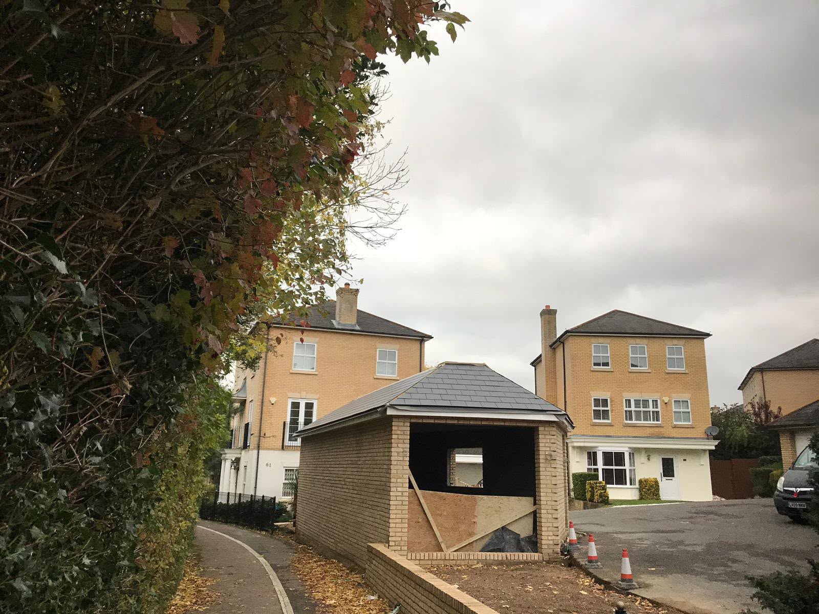 A new brick garage with a hipped slate roof and a boarded opening, seen from a leafy footpath with houses behind