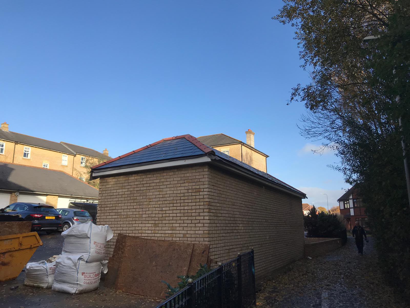 The long brick side wall of a new garage with a hipped slate roof and red ridge tiles under a blue sky