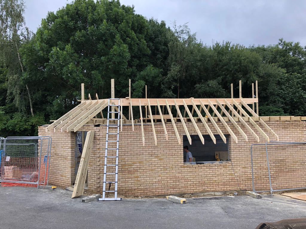 A new yellow brick garage with window and door openings and the first roof rafters in place, with a ladder against the wall