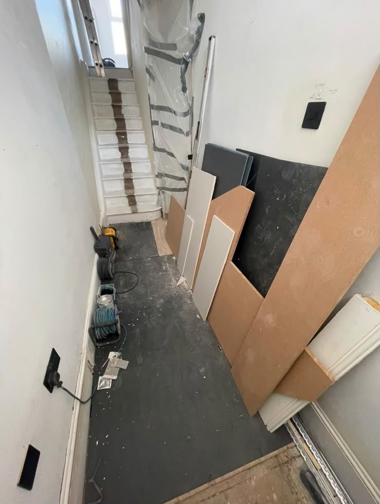 A narrow hallway stripped back to the subfloor, with the staircase protected and timber boards leaning against the wall