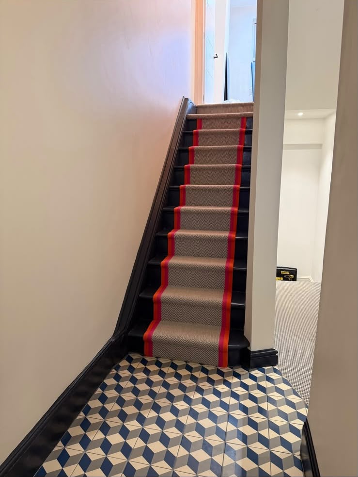 A finished hallway with geometric blue and white floor tiles and a staircase fitted with a herringbone runner edged in red and orange