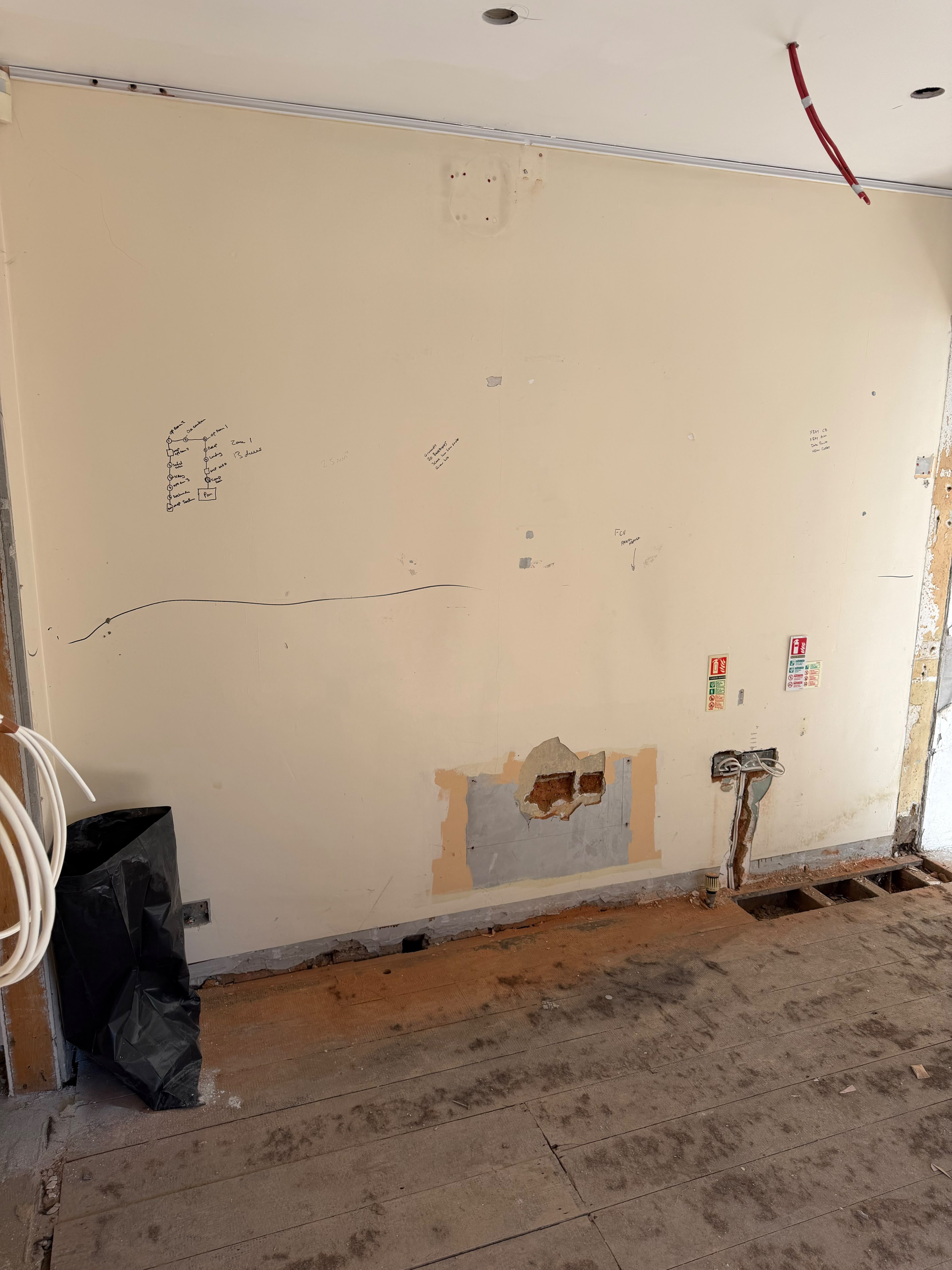 A cream painted wall with plaster broken out near its base and grey patch repairs along the bottom, above dusty floorboards