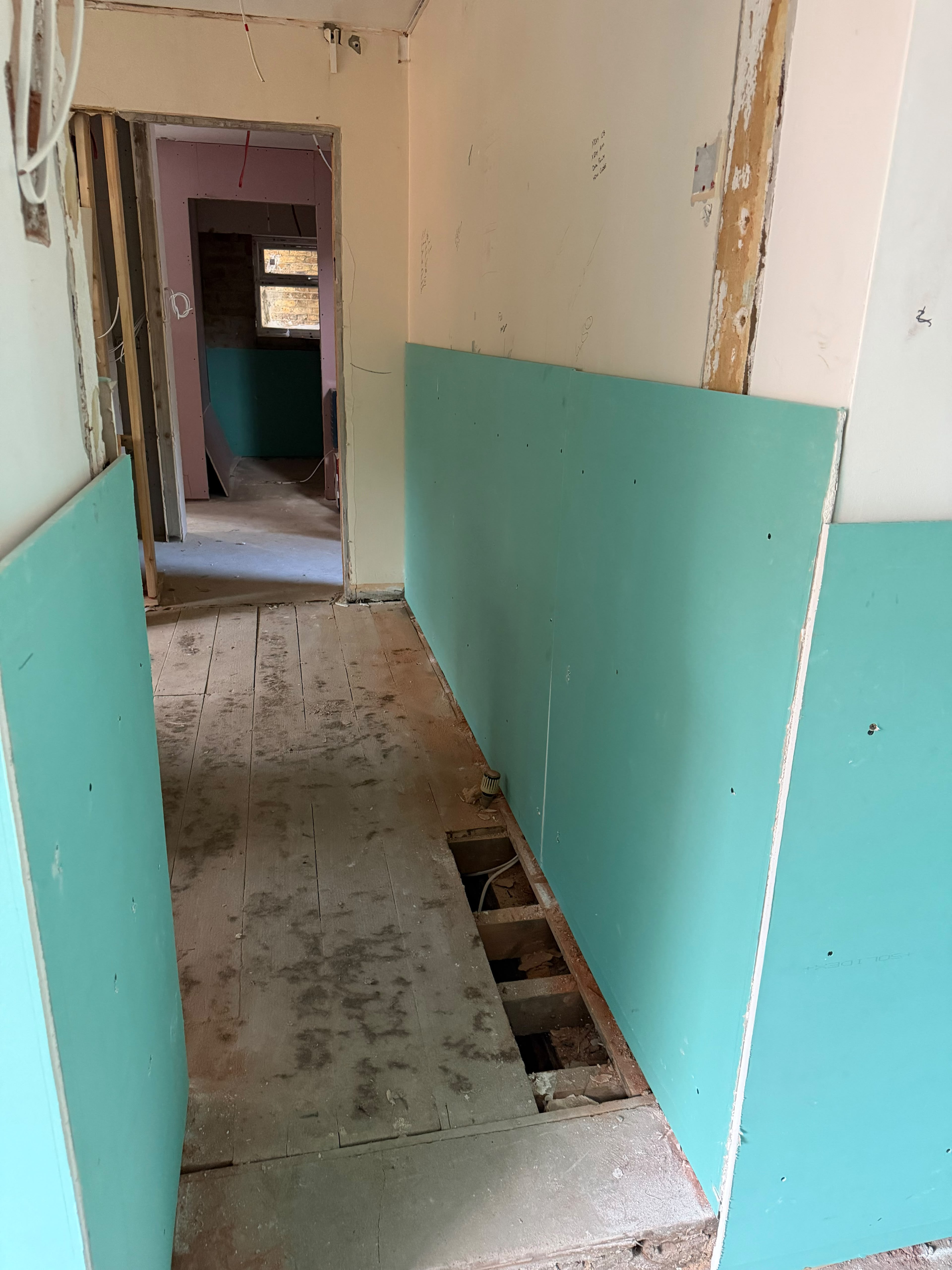 A corridor with turquoise boards on the lower walls on both sides, a section of floorboards lifted along one wall and a doorway at the end