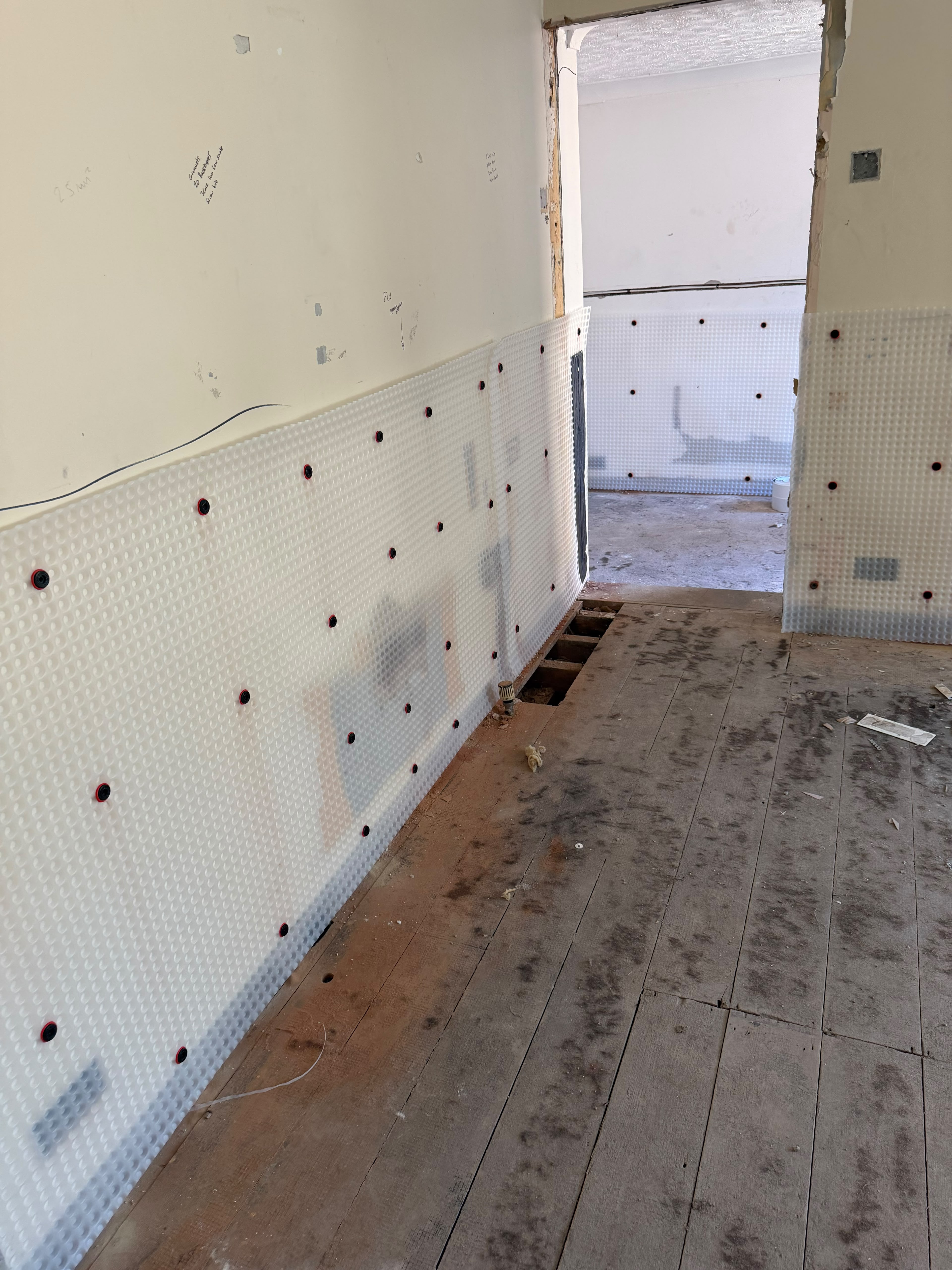 A corridor wall covered at low level in white studded membrane fixed with red-ringed plugs, leading through a doorway to a room that is also membraned