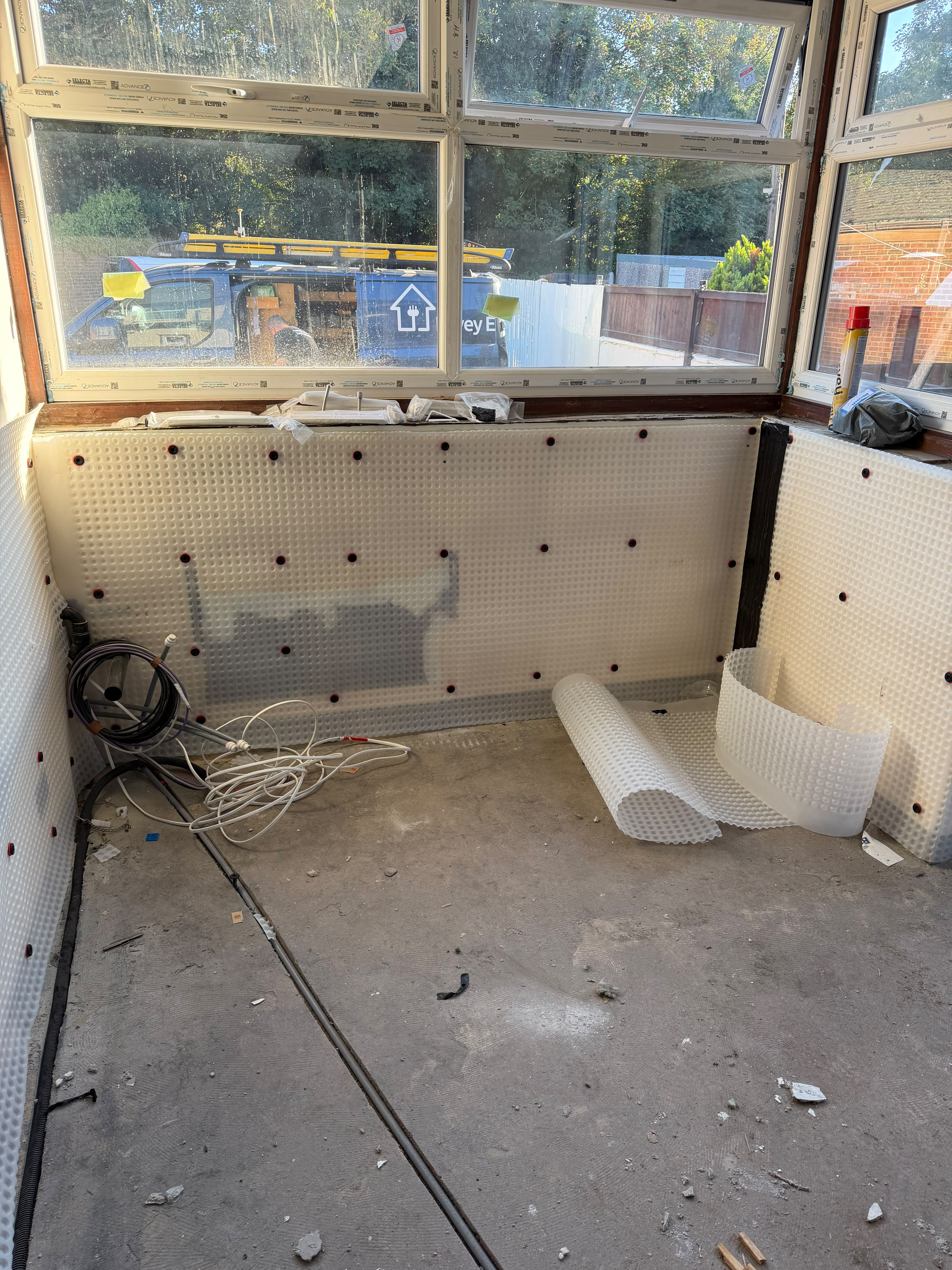 White studded cavity membrane fixed to the low walls under a bay window with red-ringed plugs, and rolls of membrane on the concrete floor