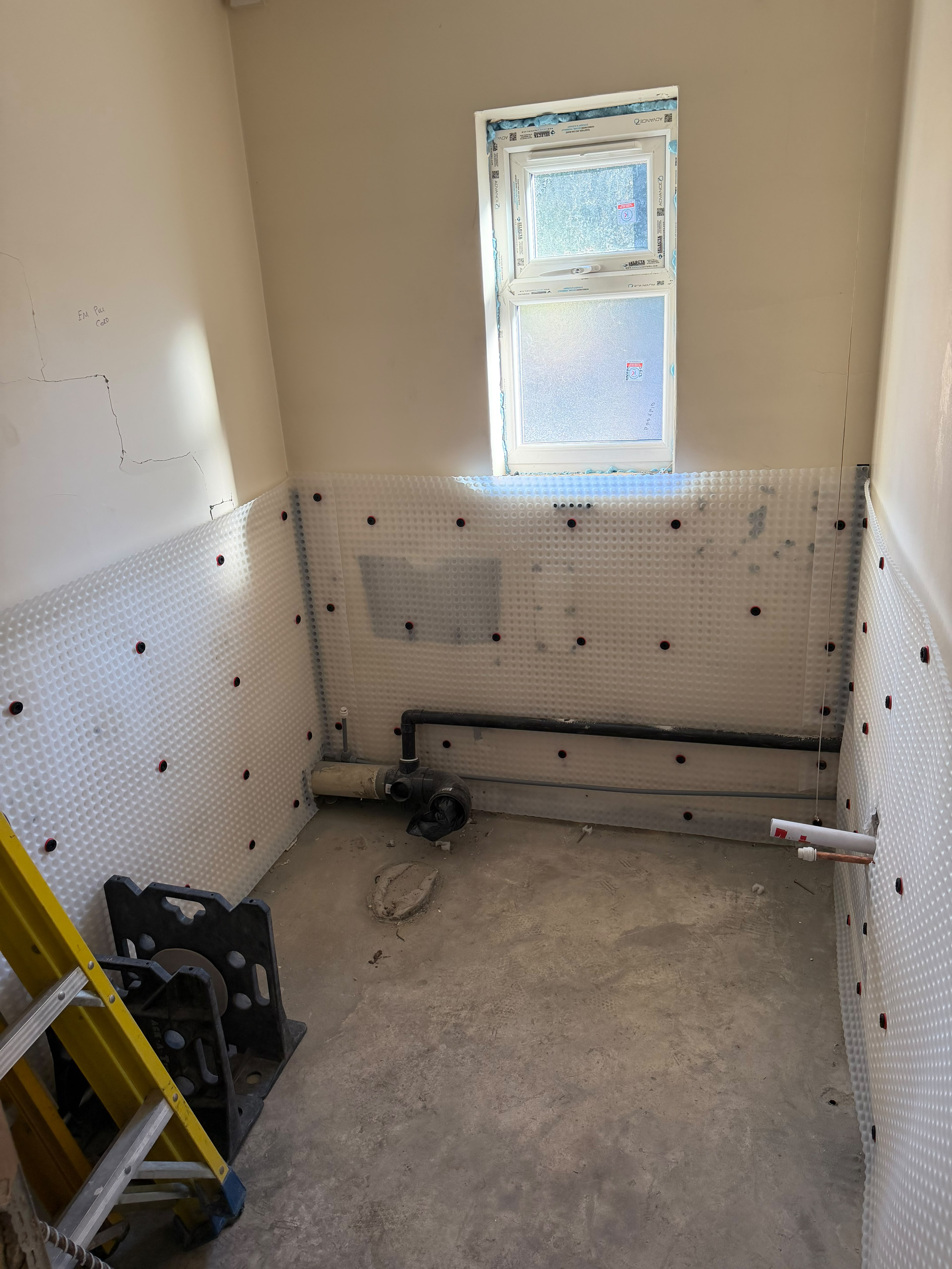 A small room with white studded membrane on the lower walls, fitted round a black waste pipe and copper supply pipes beneath a frosted window