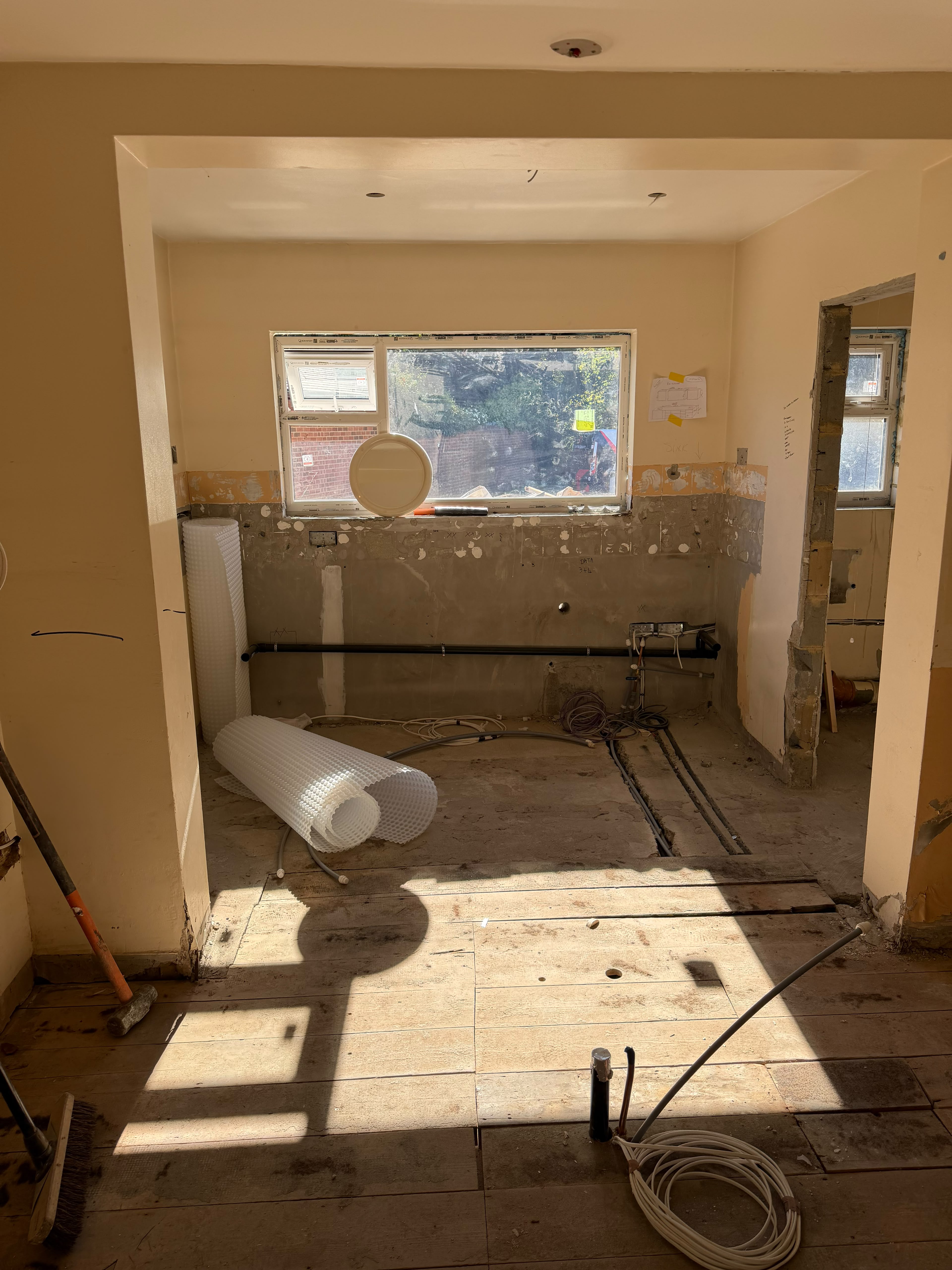 A kitchen recess under a window with the tiles removed and the wall stripped back, a pipe along the wall and rolls of white studded membrane on the floor
