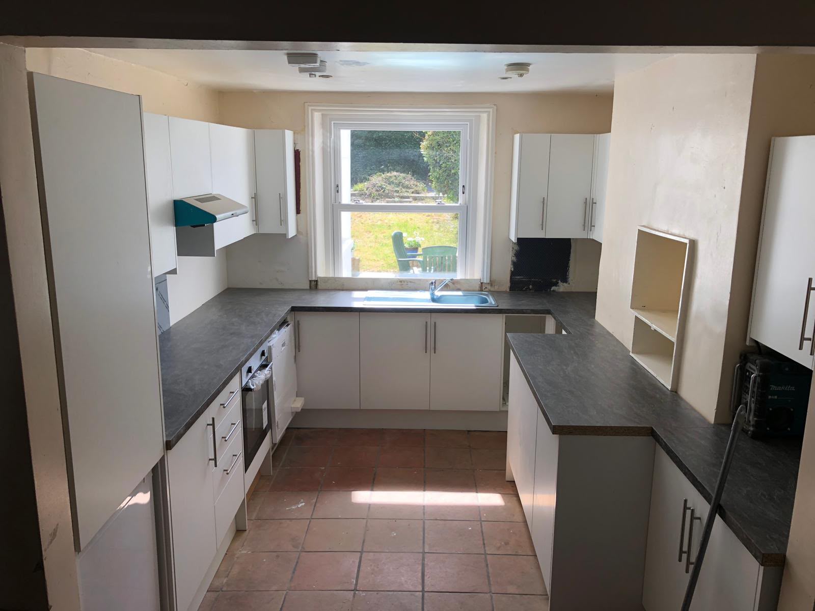 A U-shaped white kitchen with dark worktops and a sink under a sash window, still standing on the old terracotta tiled floor
