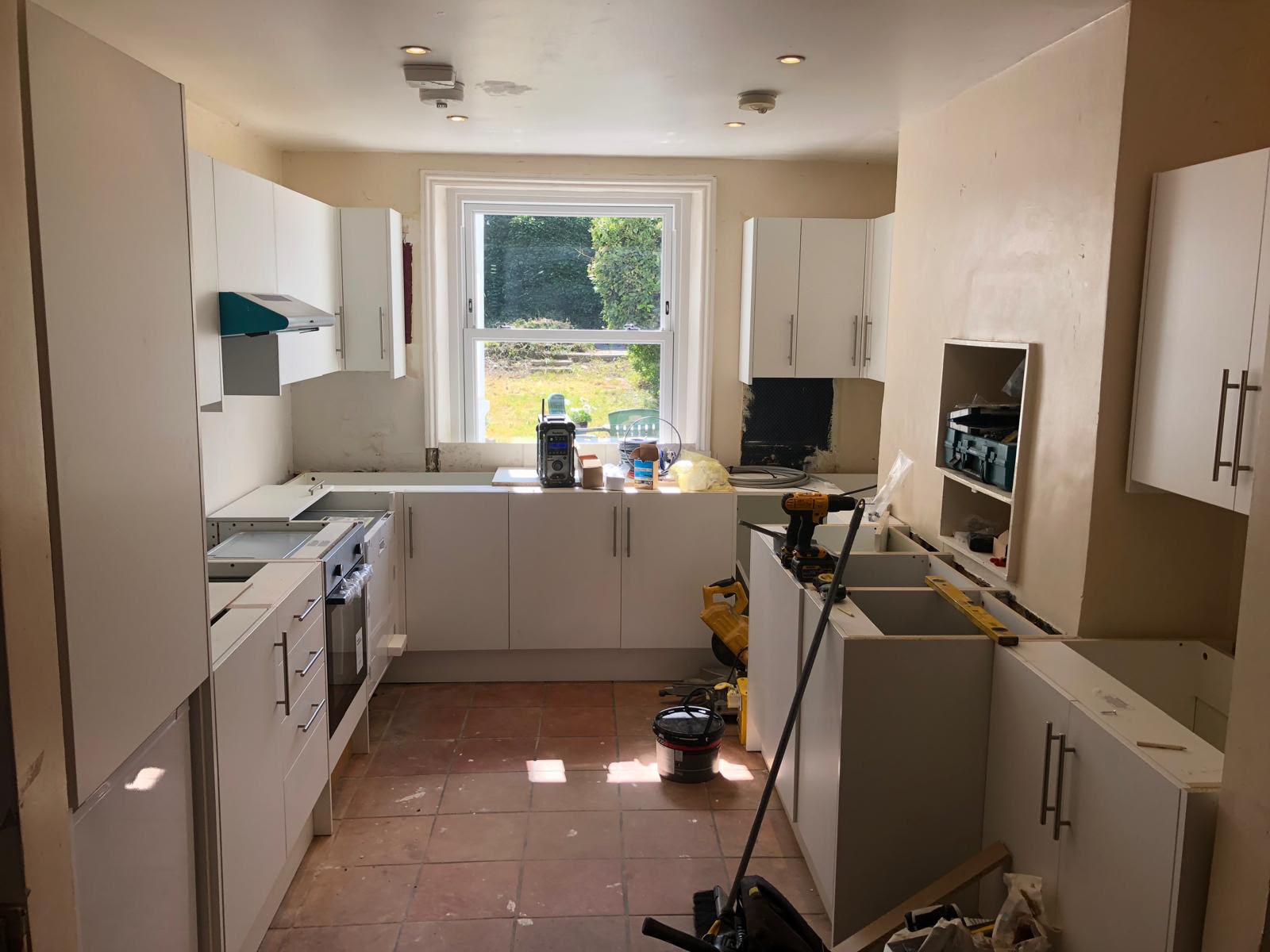 A kitchen mid-fit with white wall and base units around a sash window, worktops not yet fitted and tools on the terracotta tiled floor