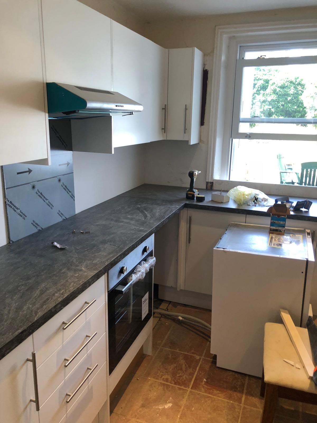 A dark stone-effect worktop fitted along white base units and drawers, with a built-in oven, an extractor hood and a drill resting on the worktop