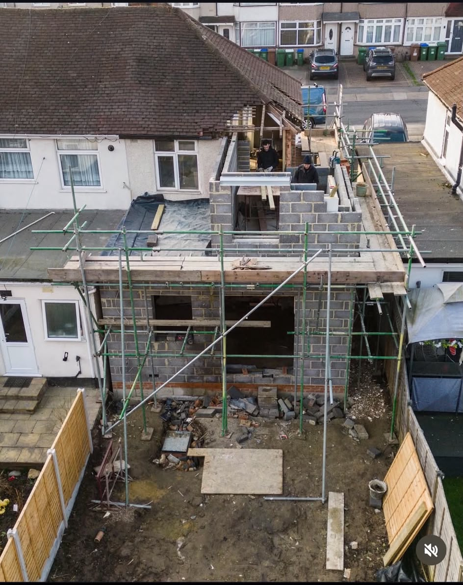 Builders laying blockwork for the first floor of a side extension, above a single-storey rear extension shell wrapped in scaffolding