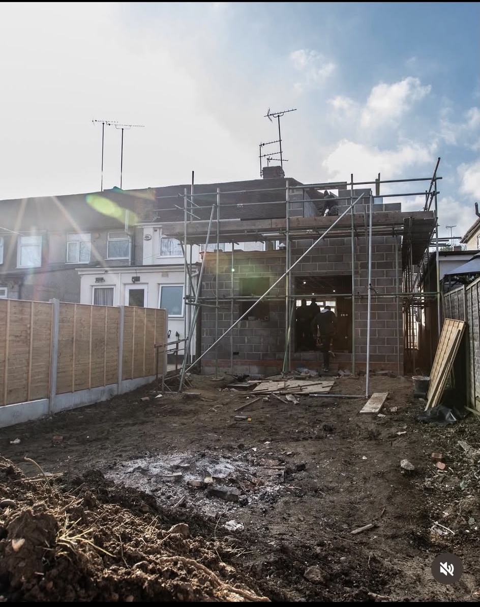 A blockwork extension shell wrapped in scaffolding, seen from a cleared garden in low sunlight