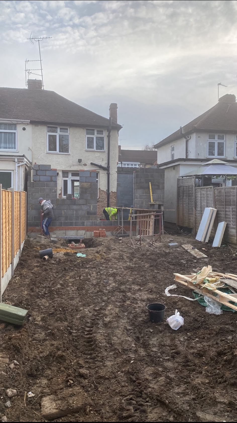 Builders raising blockwork walls for a rear extension against a semi-detached house, with a cleared muddy garden in front