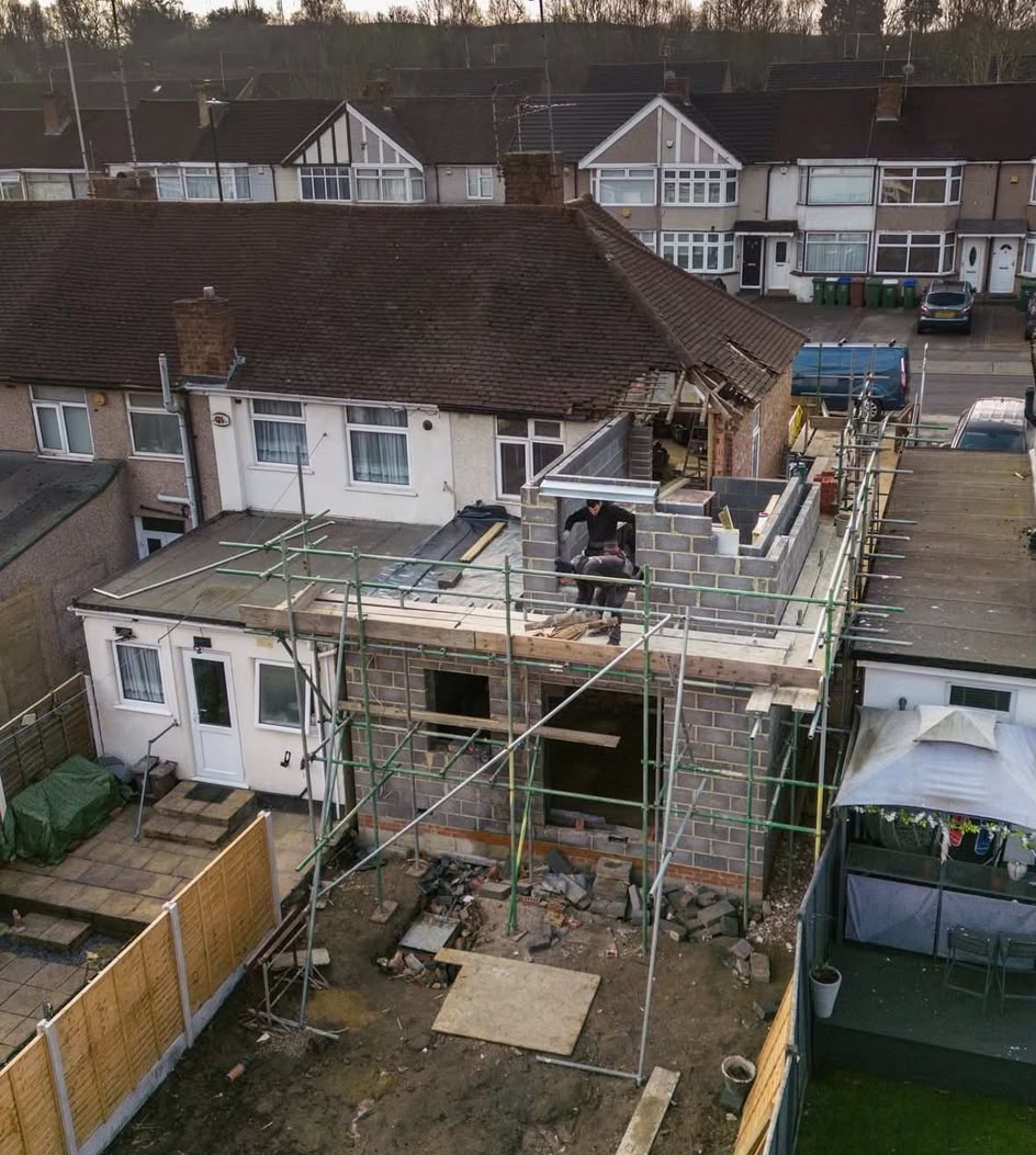 Two builders working on the first floor blockwork of a side extension next to an opened-up hipped roof