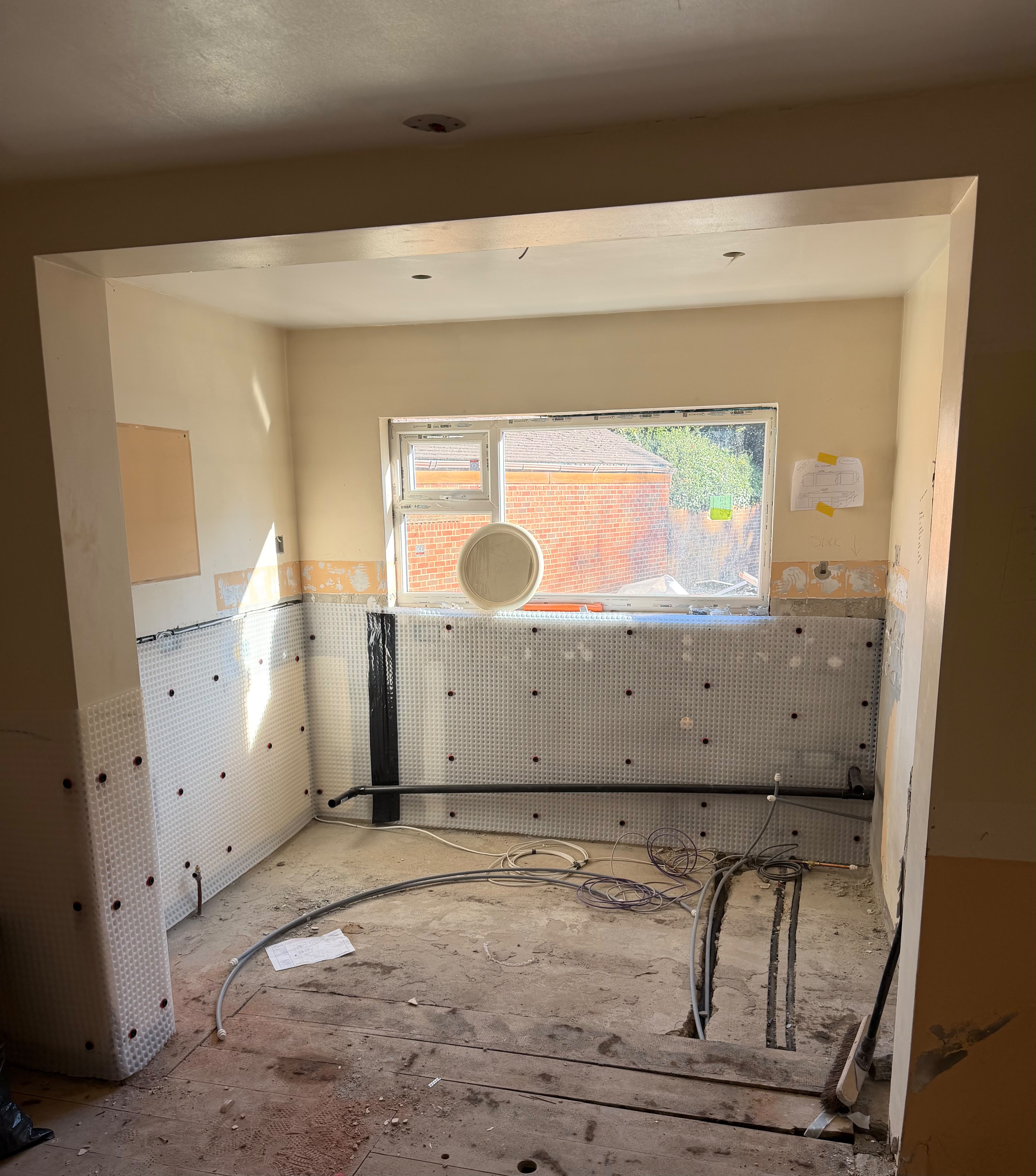 A kitchen recess with a white studded cavity wall membrane fixed to the lower walls under the window, with new pipework and cables running across the stripped floor