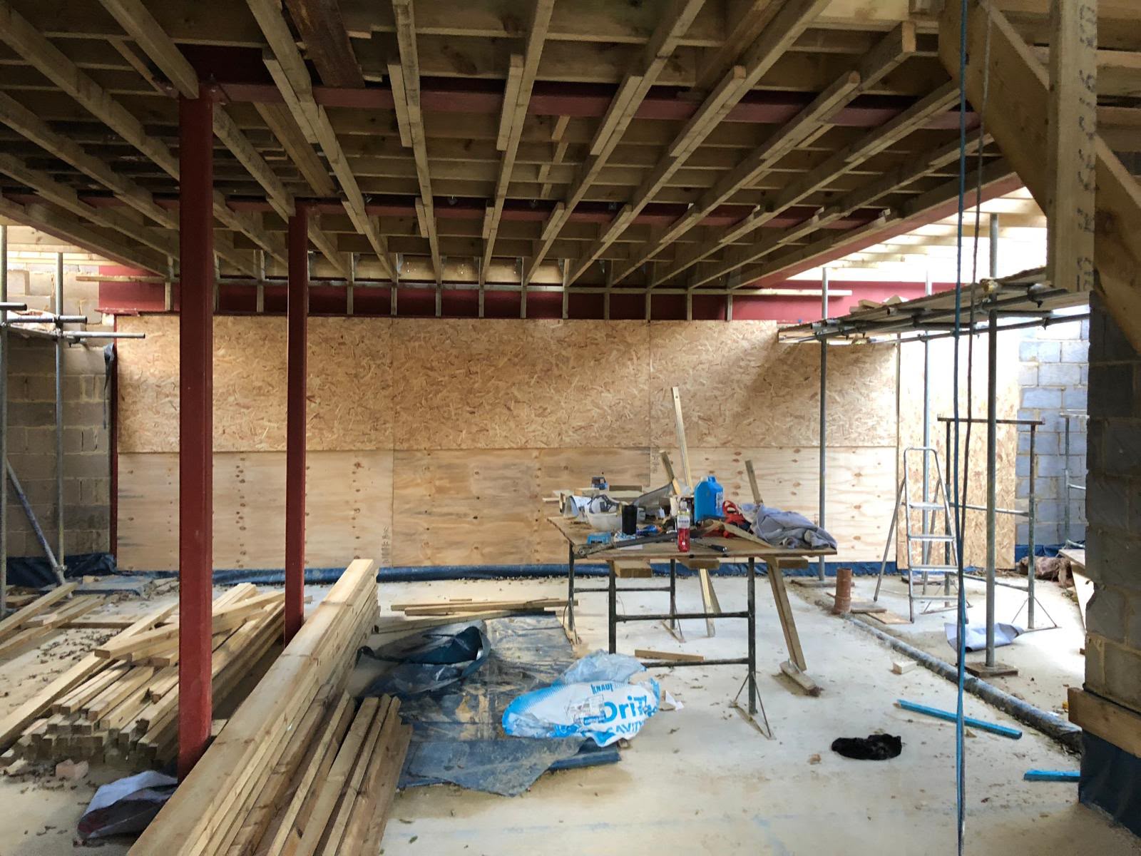 New floor joists carried on red steel beams and columns inside a house under construction, with boarded walls and timber stacked on the slab