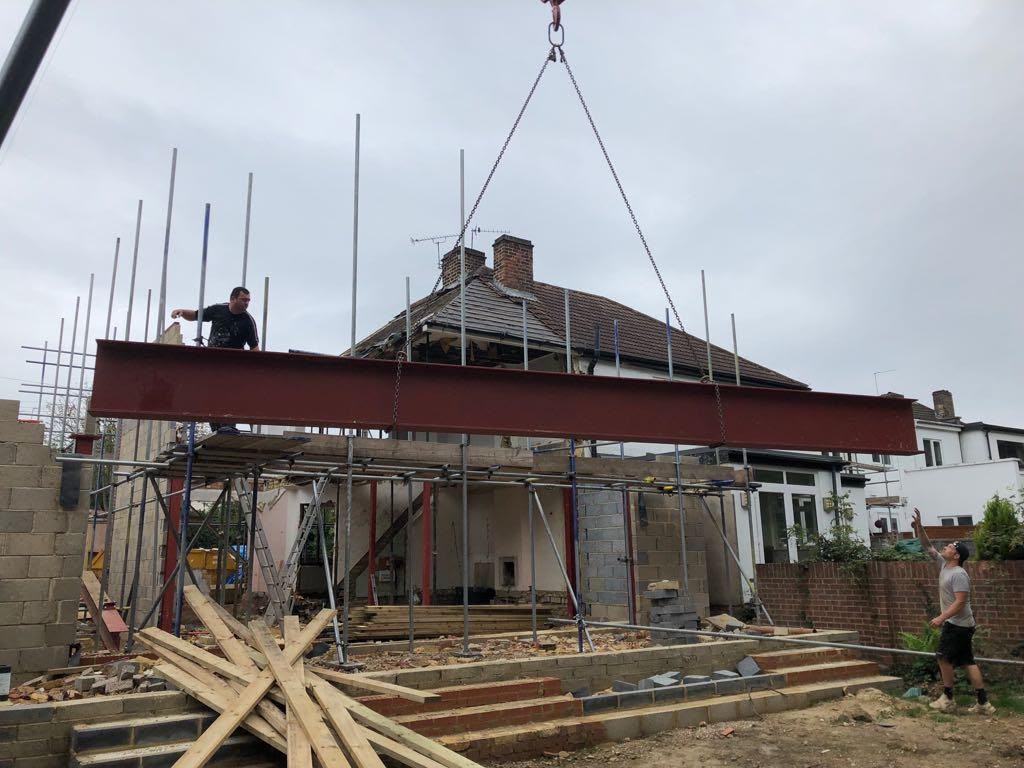 A long red steel beam being craned into place over a rear extension under construction, guided by builders on the scaffold and the ground