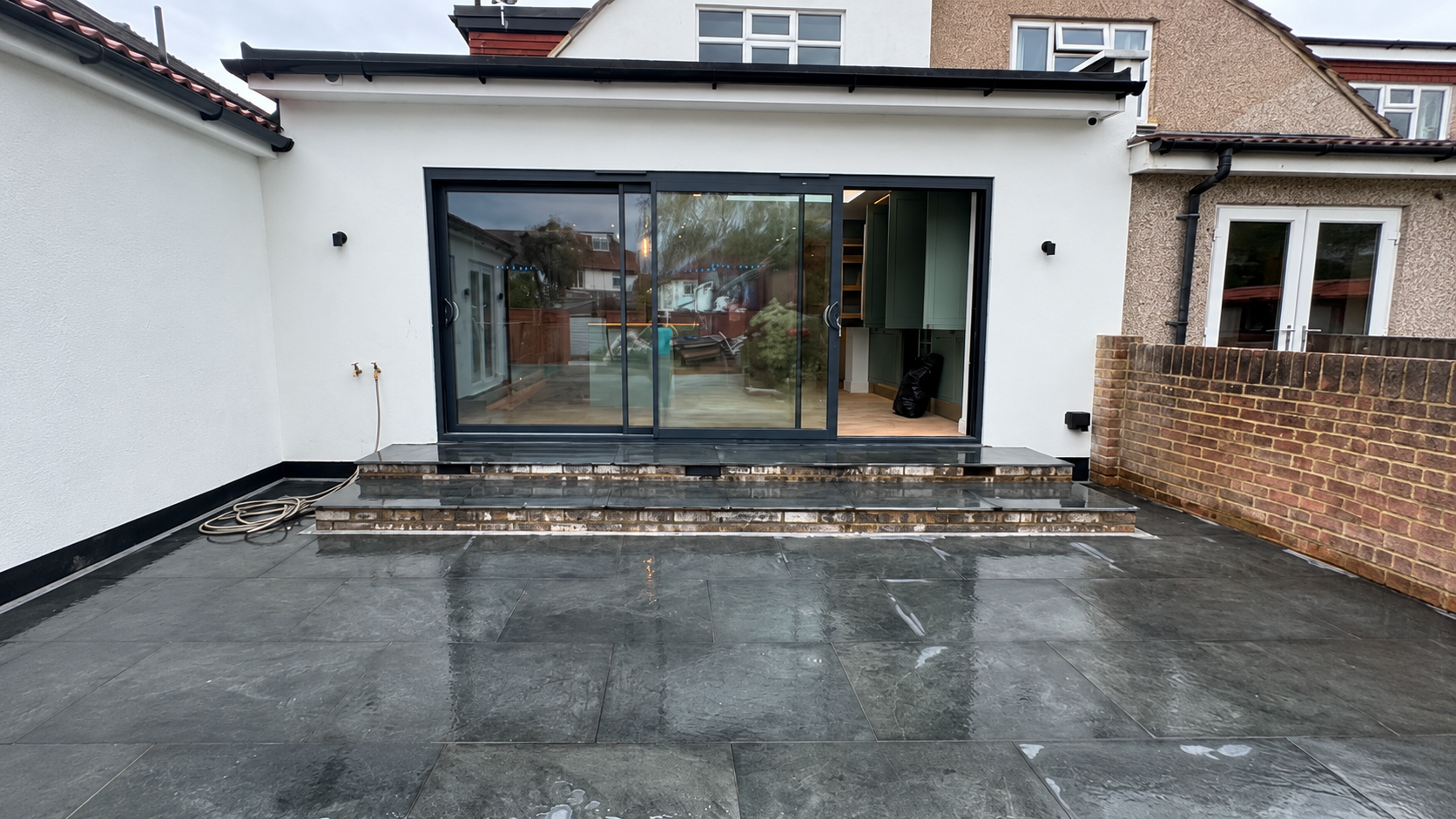 White rendered rear extension with black framed sliding doors, brick steps and a dark paved patio